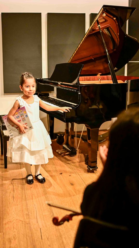 oung girl in a white dress standing proudly next to a grand piano after her spring recital.